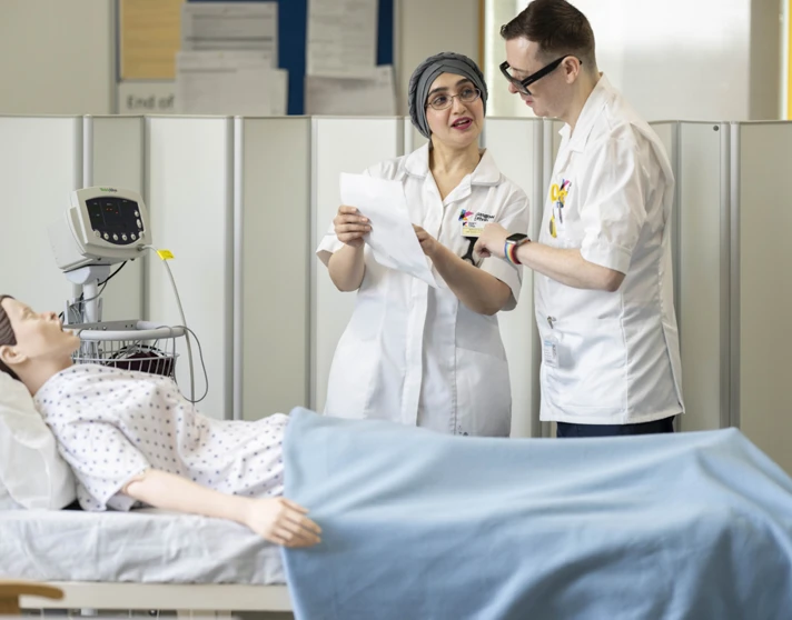 Two care students discuss notes at a hospital bed during a training exercise Two care students discuss notes at a hospital bed during a training exercise